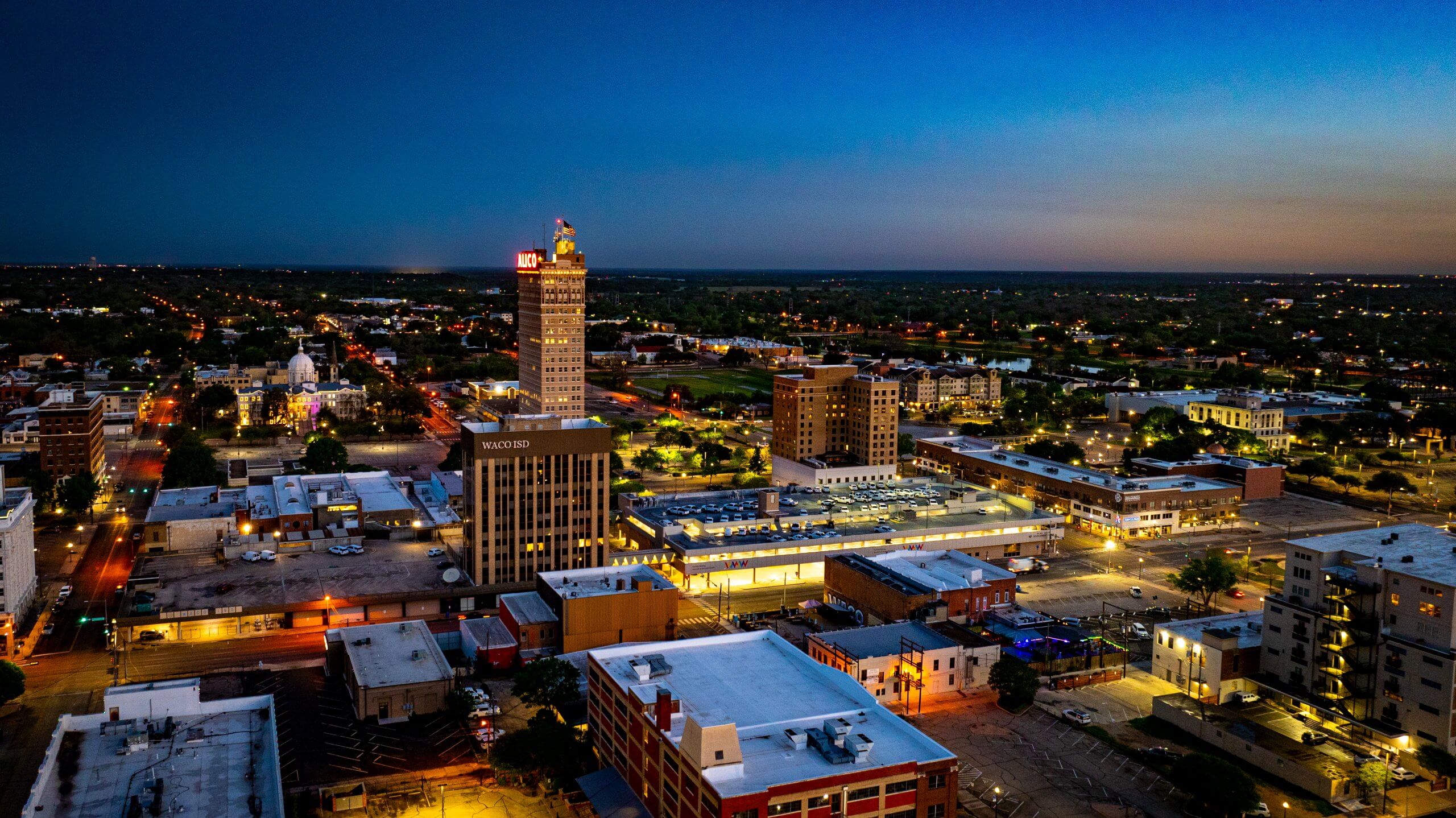 An aerial view of the Waco skyline at dusk with illuminated buildings in McLennan County, Texas.
