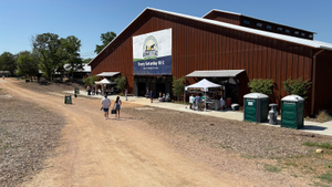 People walk on a dirt path toward a large brown barn with market stalls in McLennan County, Texas.