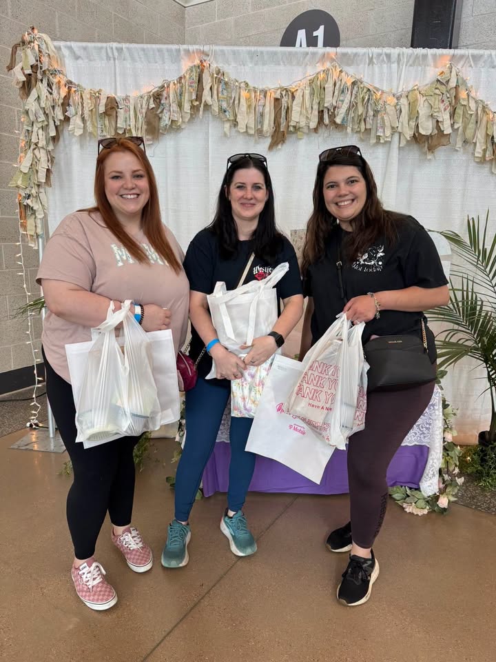 Three women smiling while holding shopping bags in front of a decorated backdrop in McLennan County, Texas.