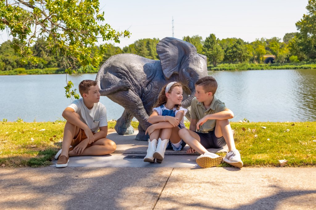 Three children sitting on the ground next to a bronze elephant statue by a lake in McLennan County, Texas.