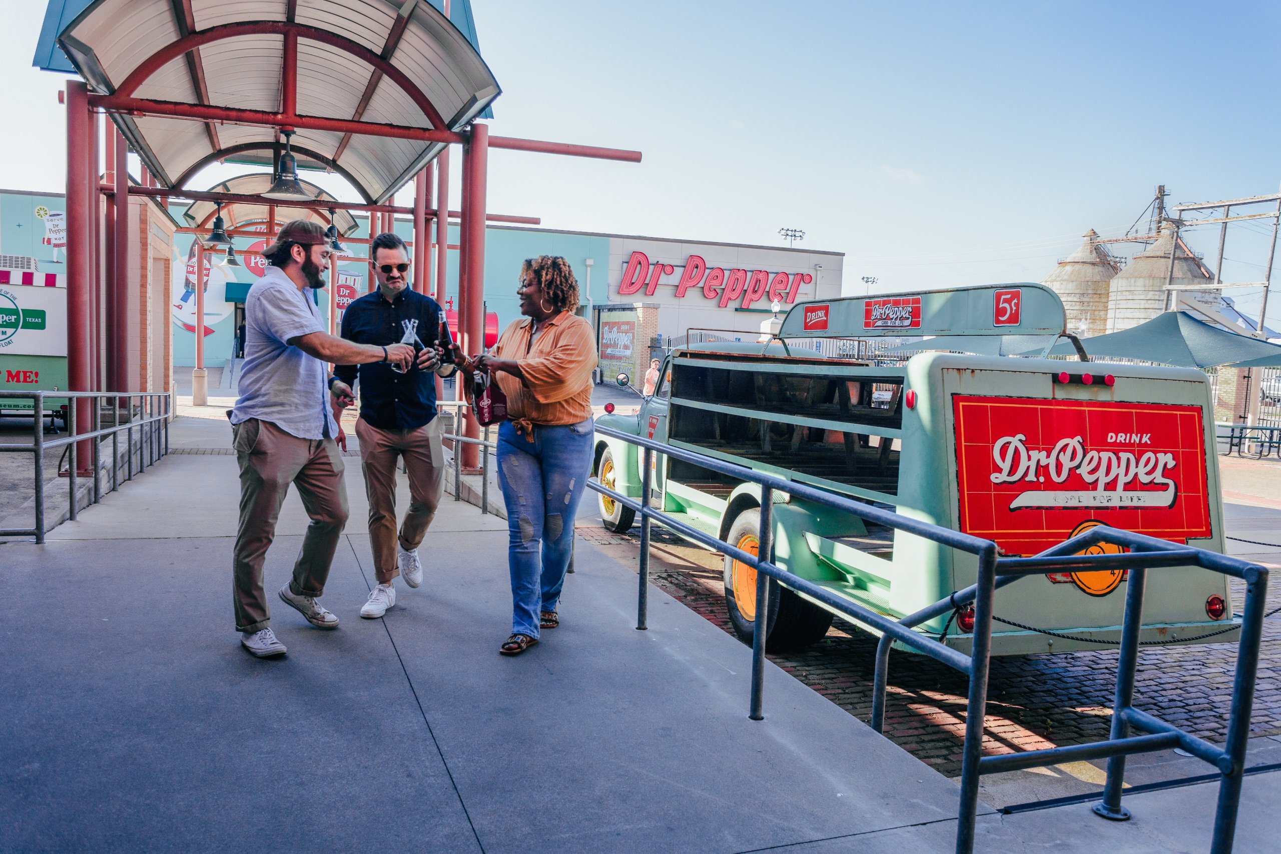 Three friends walking and toasting with glass bottles near a vintage Dr. Pepper truck in McLennan County, Texas.