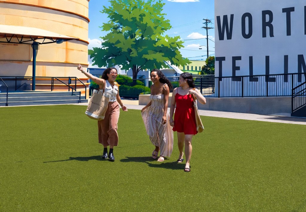 Three women walking and laughing on a green lawn near a large mural in McLennan County, Texas.