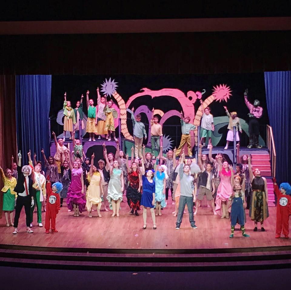 A large group of children in colorful costumes performing on a theater stage in McLennan County, Texas.