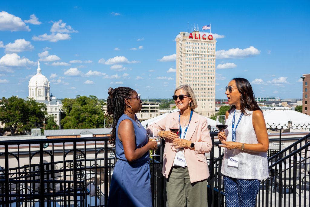 Three women socialize with wine on a rooftop terrace overlooking a city skyline in McLennan County, Texas.