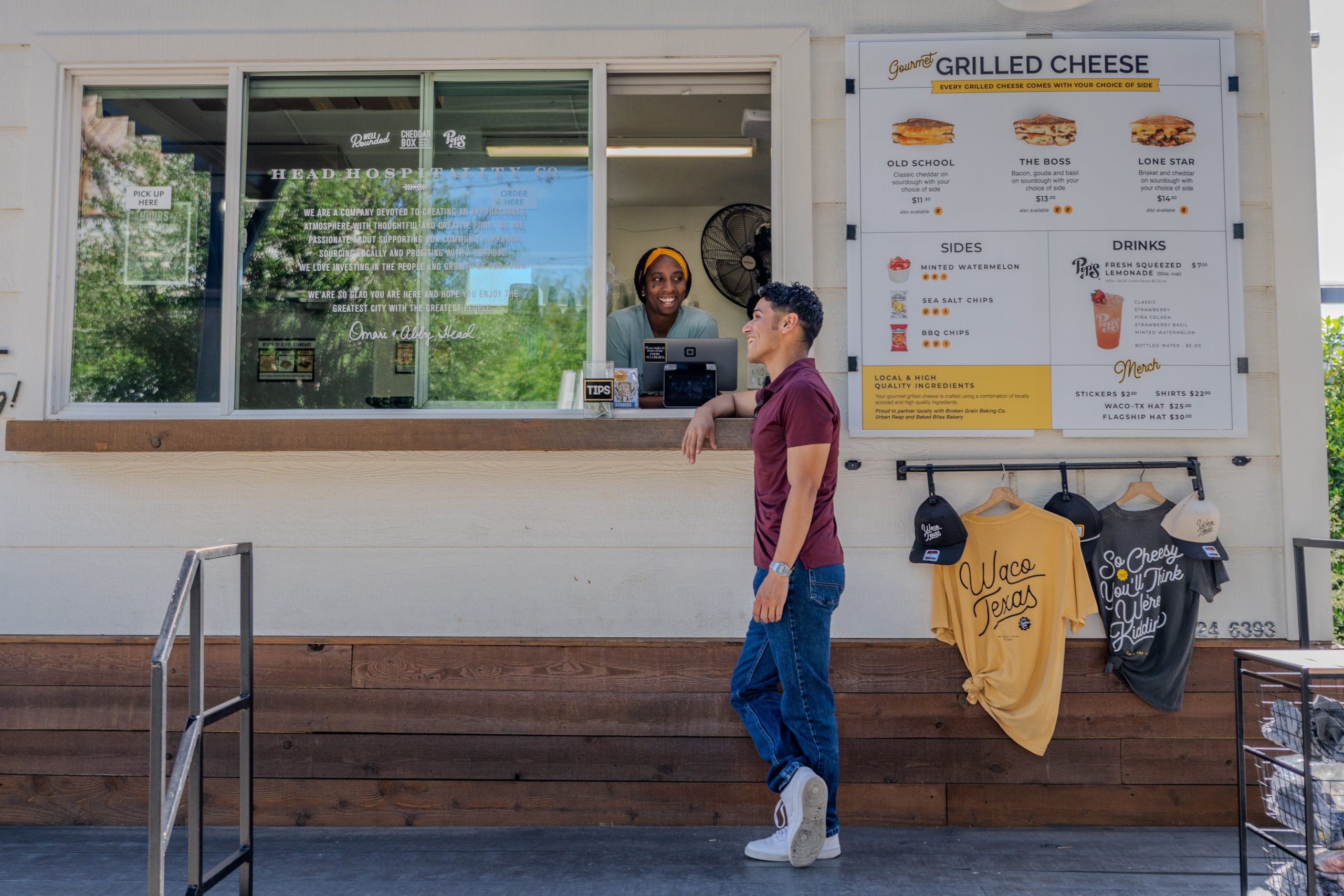A man orders food at a grilled cheese stand window from a smiling employee in McLennan County, Texas.