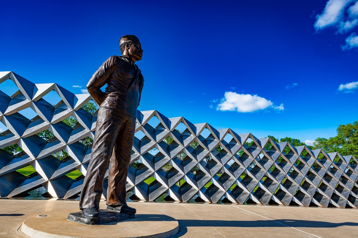 A bronze statue of a sailor standing before a geometric metal wall under blue sky in McLennan County, Texas.