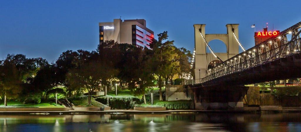 A suspension bridge and city buildings illuminated at night by a river in McLennan County, Texas.