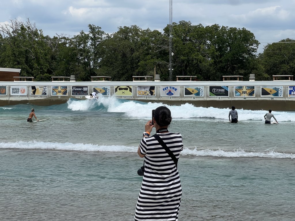 A person in a striped dress photographs surfers at an artificial wave pool in McLennan County, Texas.