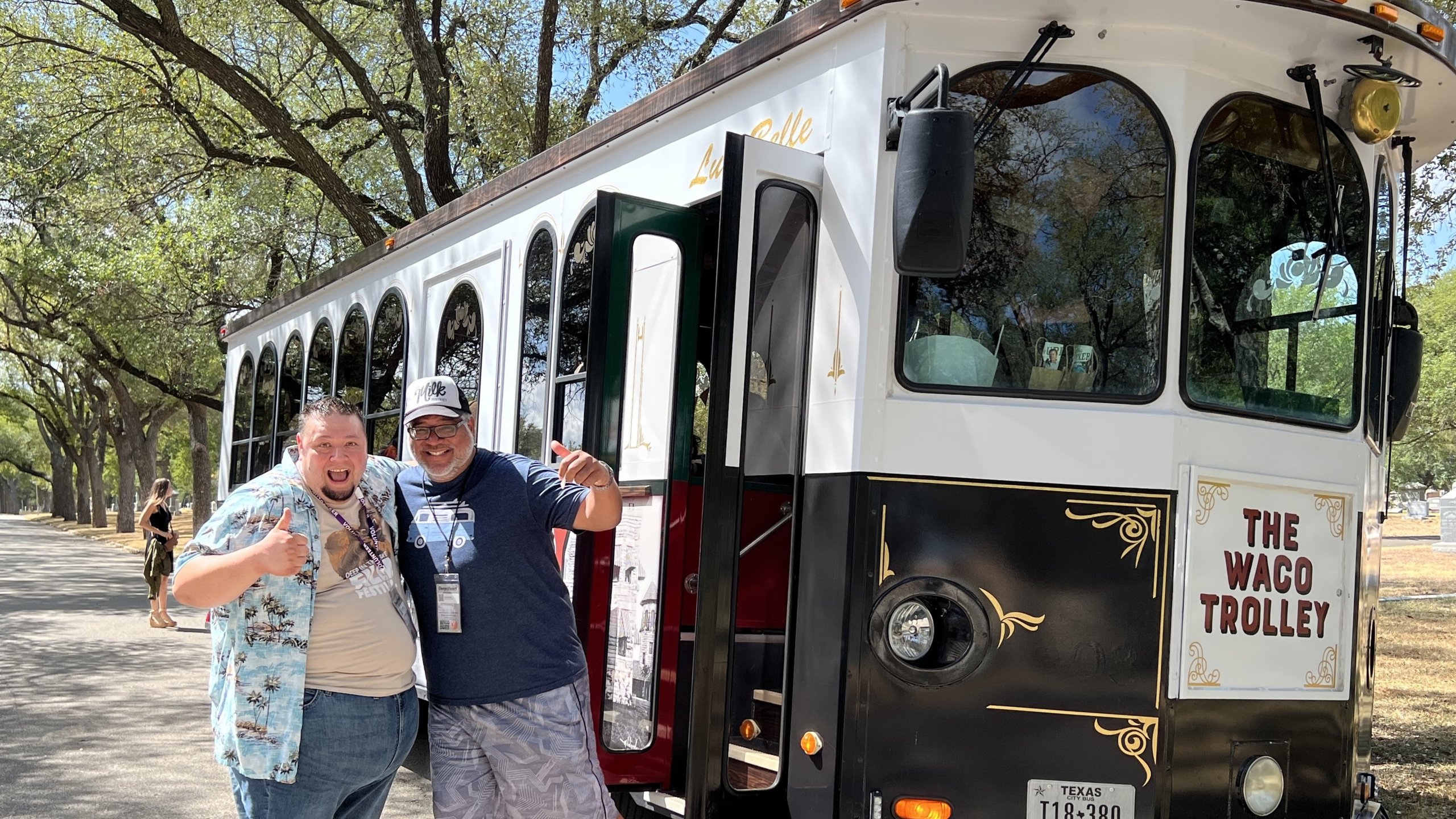 Two men smiling and giving thumbs up in front of a Waco Trolley bus in McLennan County, Texas.