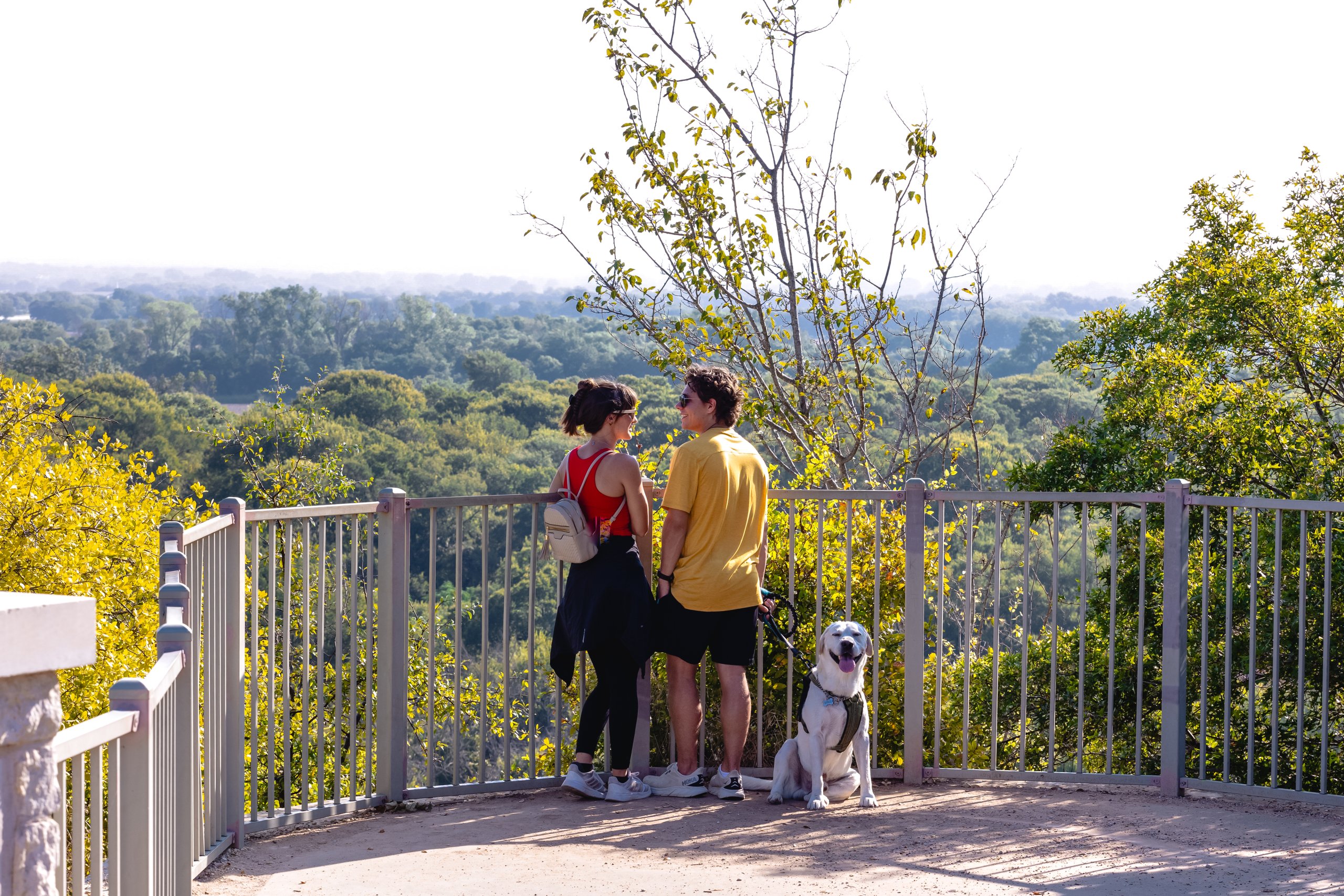 A couple and their white dog stand on an overlook viewing a forest in McLennan County, Texas.