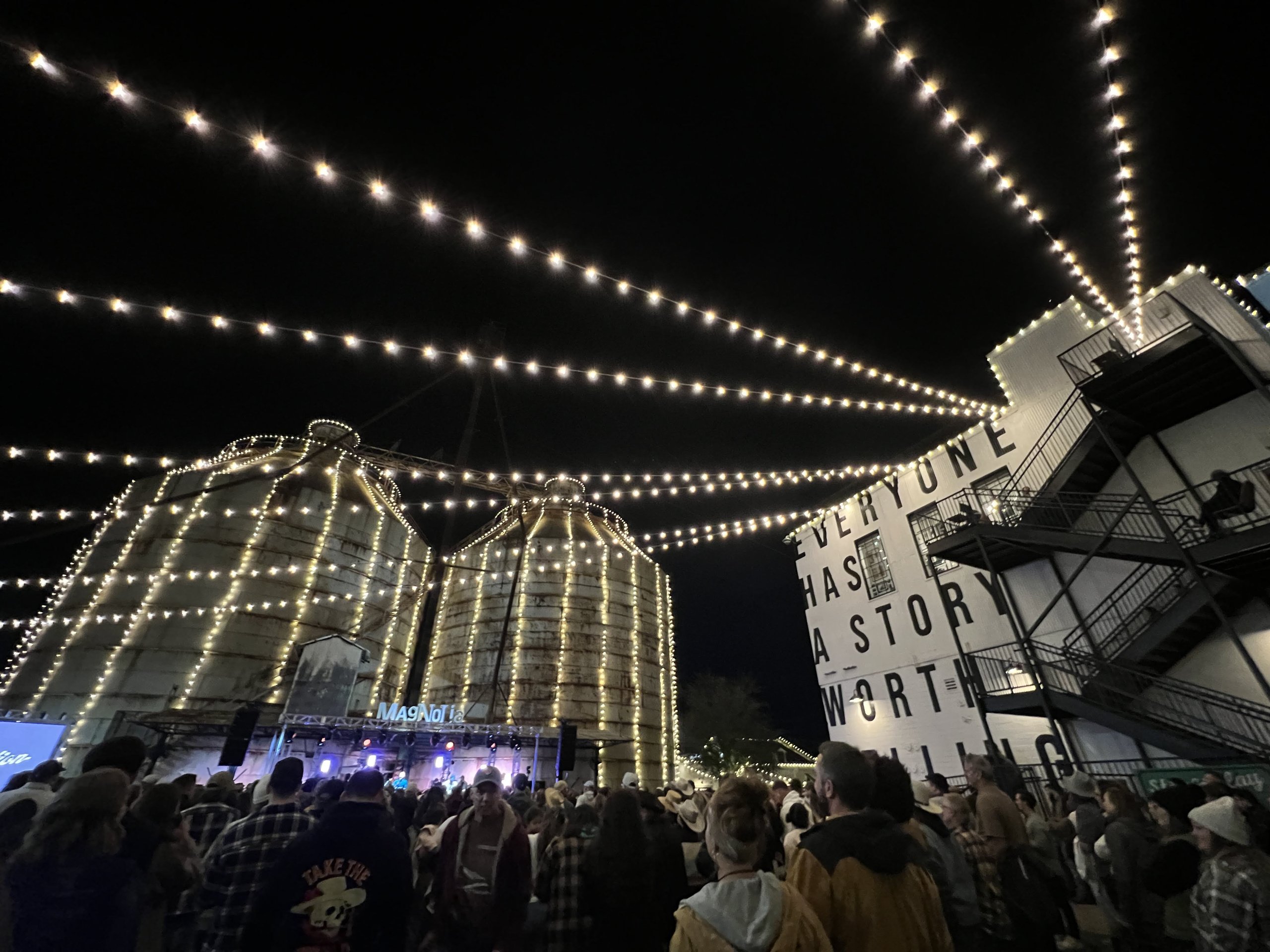 A crowd gathers at night beneath string lights near large silos and a building in McLennan County, Texas.