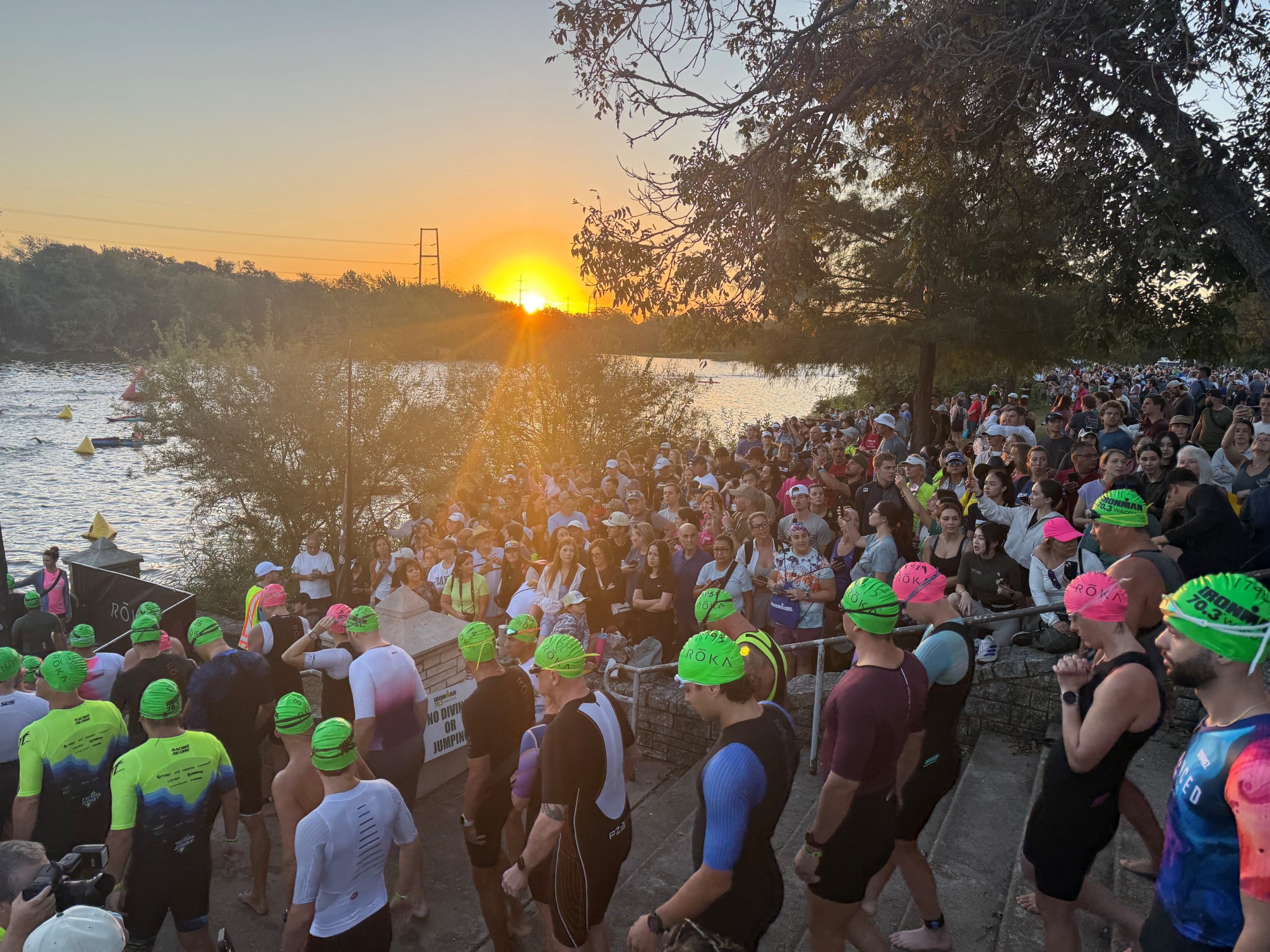 Swimmers in wetsuits and colorful caps gather at a riverbank during a sunrise triathlon in McLennan County, Texas.