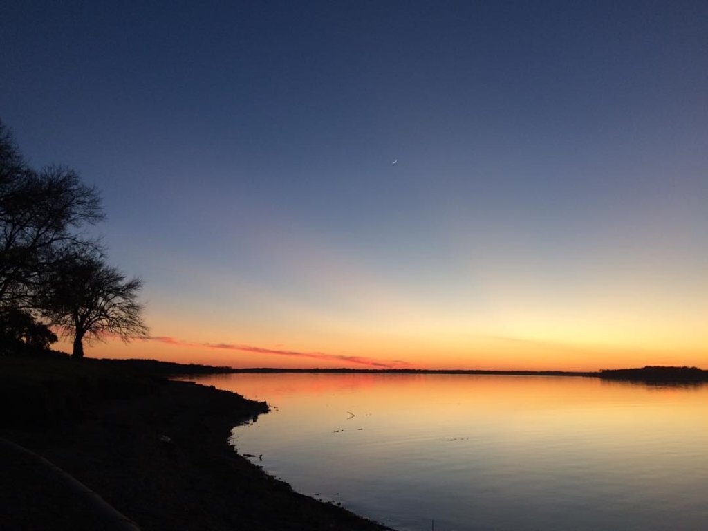 A serene lake at sunset with a crescent moon in the sky in McLennan County, Texas.