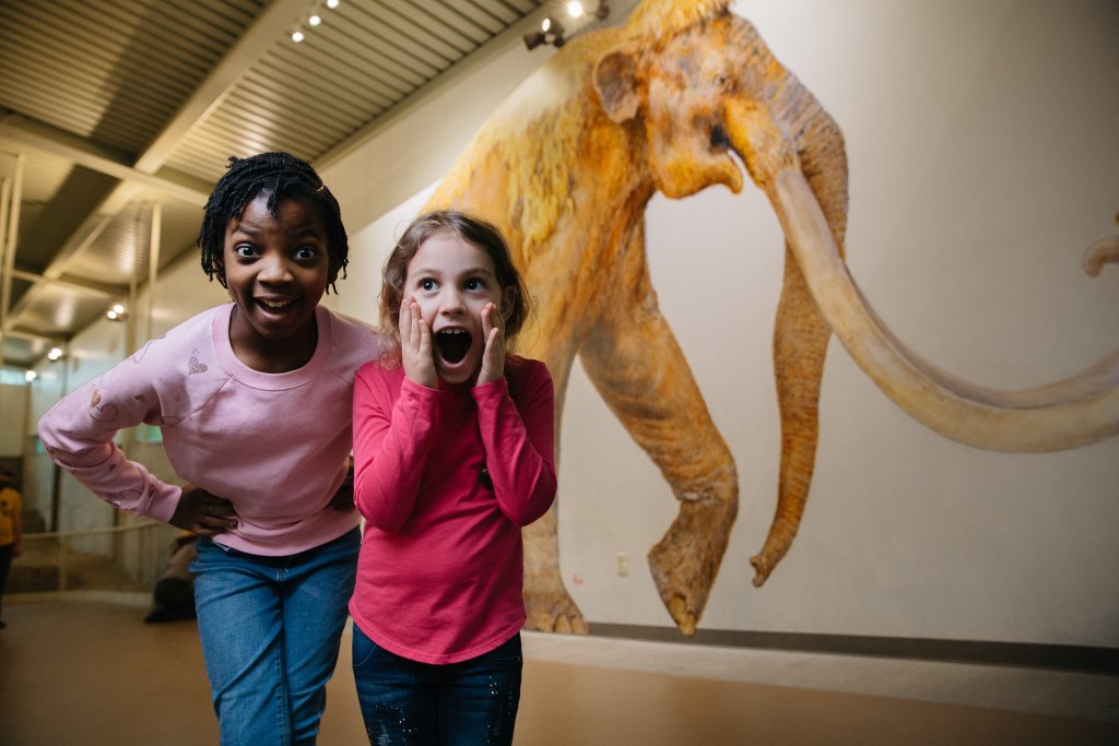 Two young girls looking excited in front of a large mammoth mural in McLennan County, Texas.