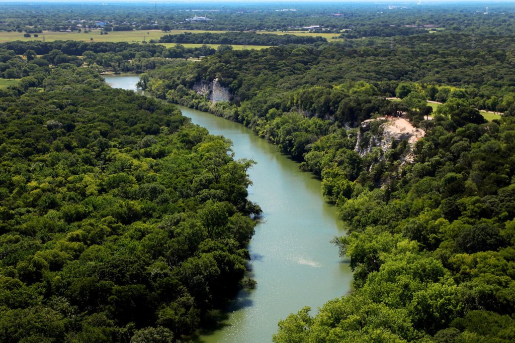 A winding river flows through a dense green forest with rocky limestone cliffs in McLennan County, Texas.