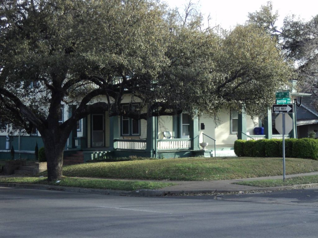A yellow house with a green porch sits behind a large tree on a corner in McLennan County, Texas.