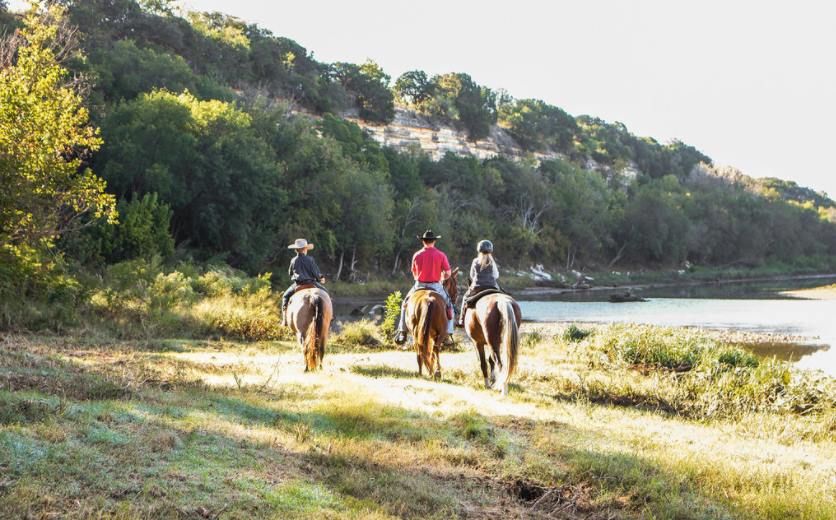 Three people ride horses along a riverbank near a wooded hillside in McLennan County, Texas.