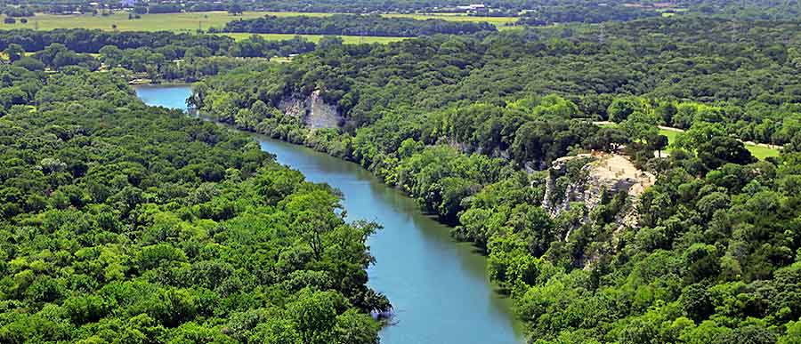 A winding river flows through a lush green forest with rocky cliffs in McLennan County, Texas.