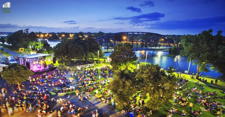 A crowd gathers in a park at dusk for an outdoor concert by a river in McLennan County, Texas.