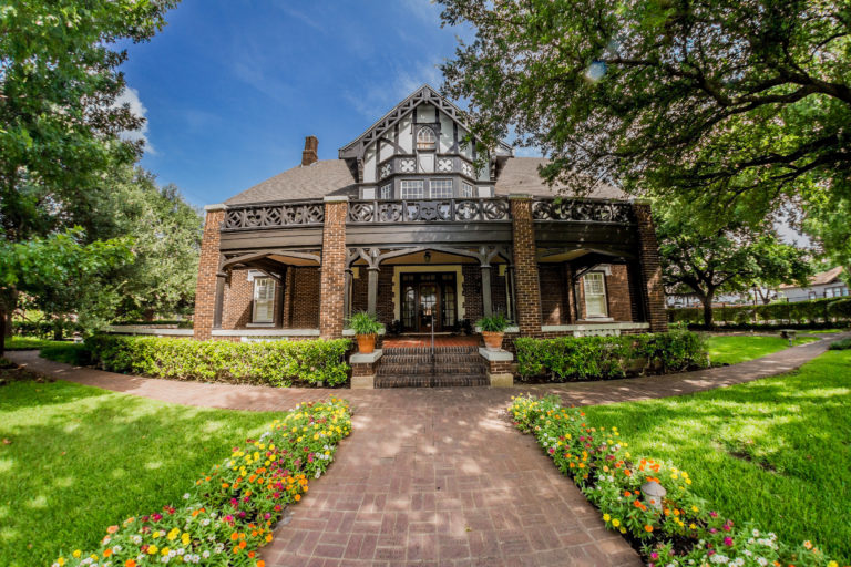A historic brick house with a large porch and a flower-lined brick walkway in McLennan County, Texas.
