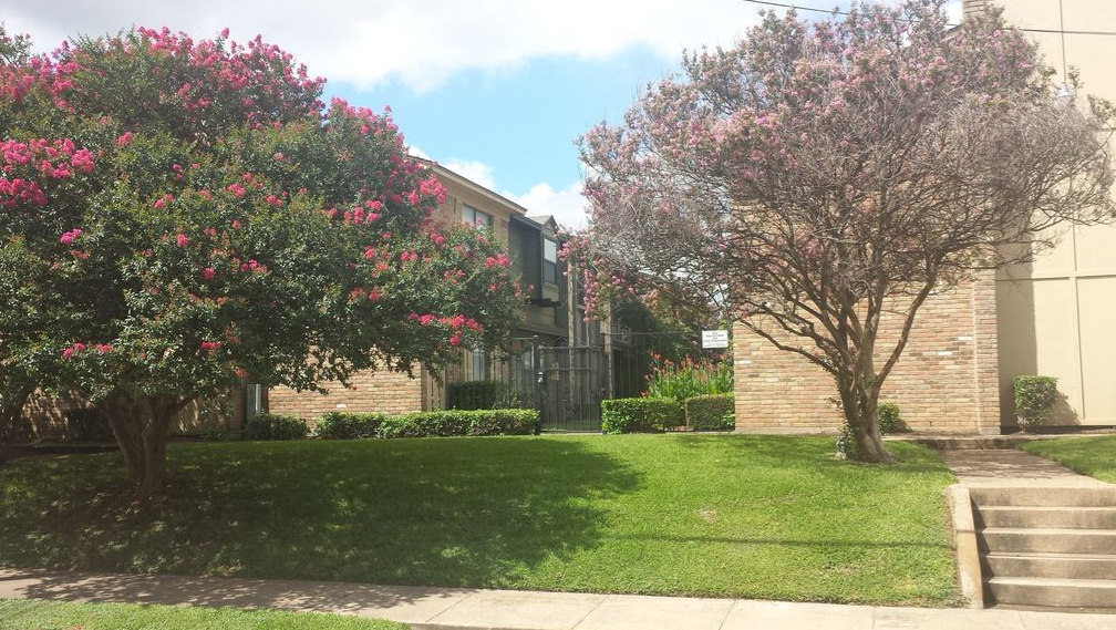 A brick apartment building surrounded by blooming crepe myrtle trees and a green lawn in McLennan County, Texas.