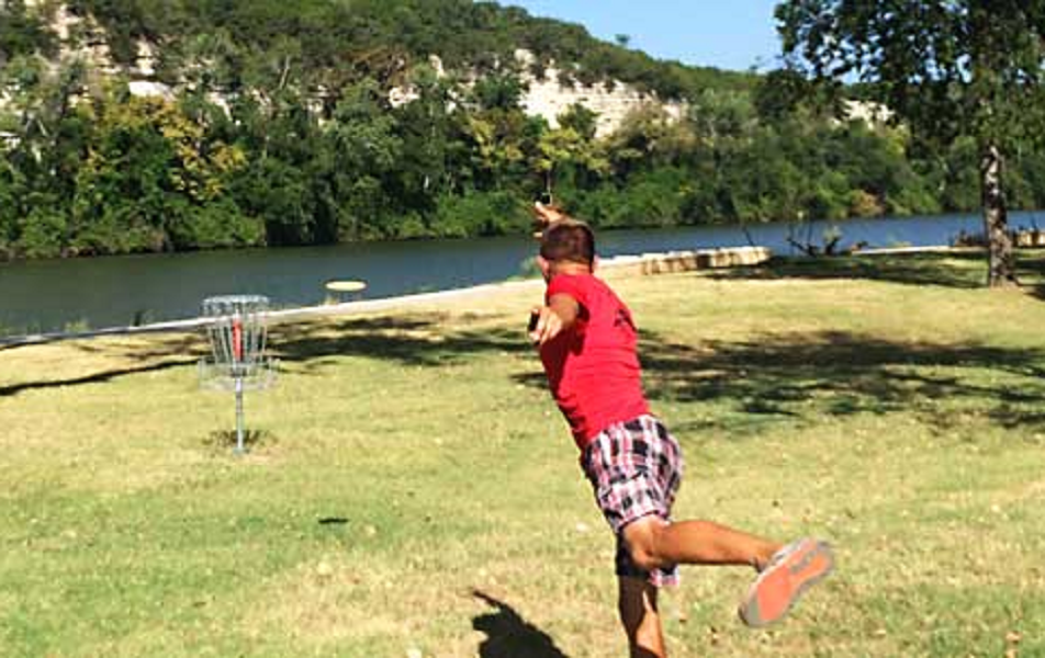 A man in a red shirt throws a disc toward a disc golf basket in McLennan County, Texas.