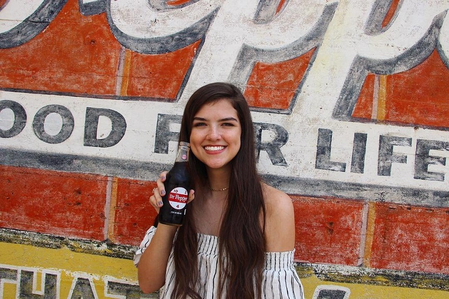 A smiling young woman holding a glass bottle of Dr Pepper in front of a wall in McLennan County, Texas.