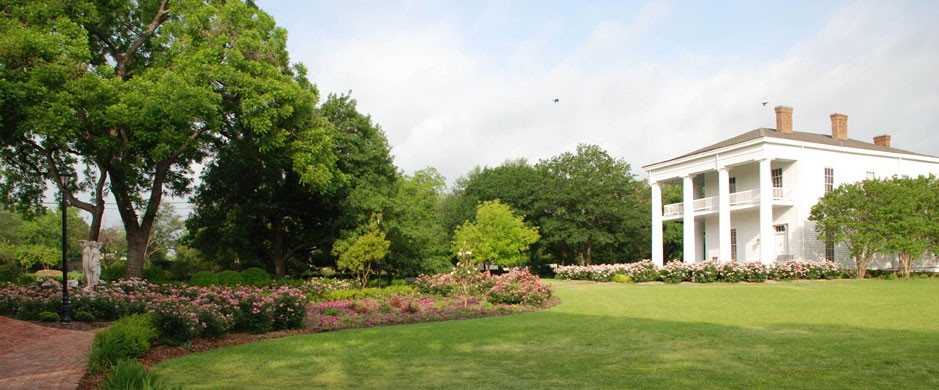 A large white antebellum-style house sits beside a lush garden and green lawn in McLennan County, Texas.