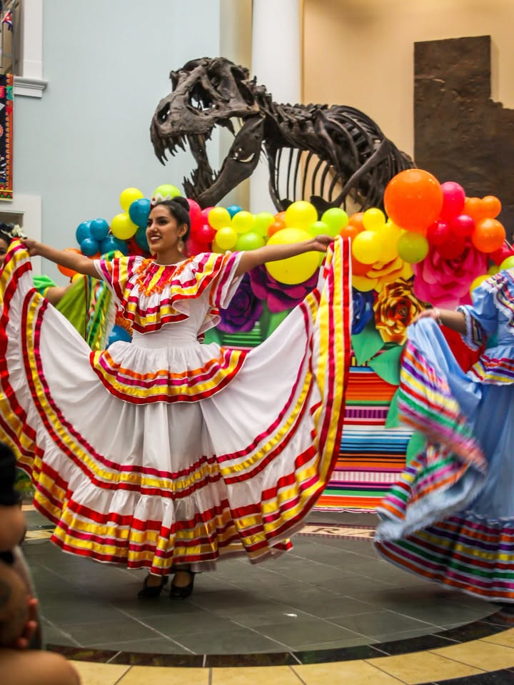 A woman in a traditional colorful Mexican dress dancing in front of a dinosaur skeleton in McLennan County, Texas.