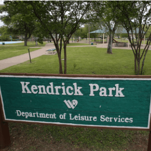 A green sign for Kendrick Park stands in front of a grassy public park in McLennan County, Texas.