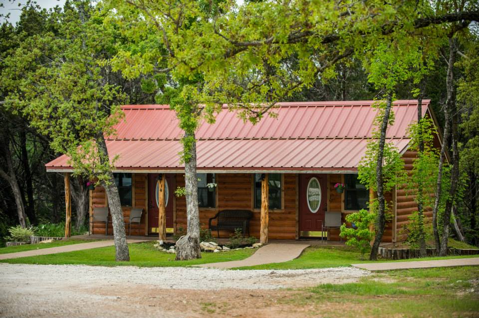 A rustic log cabin with a red metal roof surrounded by lush green trees in McLennan County, Texas.