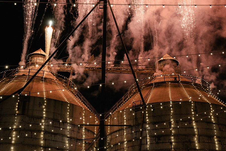 Fireworks explode above two large industrial silos decorated with strings of lights at night in McLennan County, Texas.