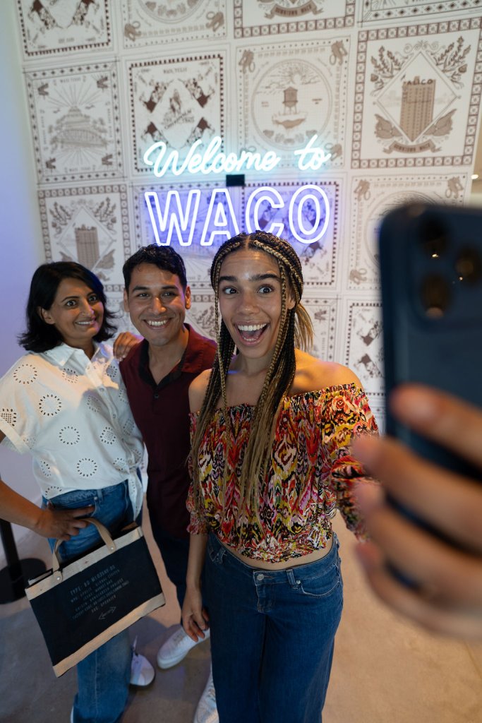 Three friends smiling while taking a selfie in front of a Waco neon sign in McLennan County, Texas.