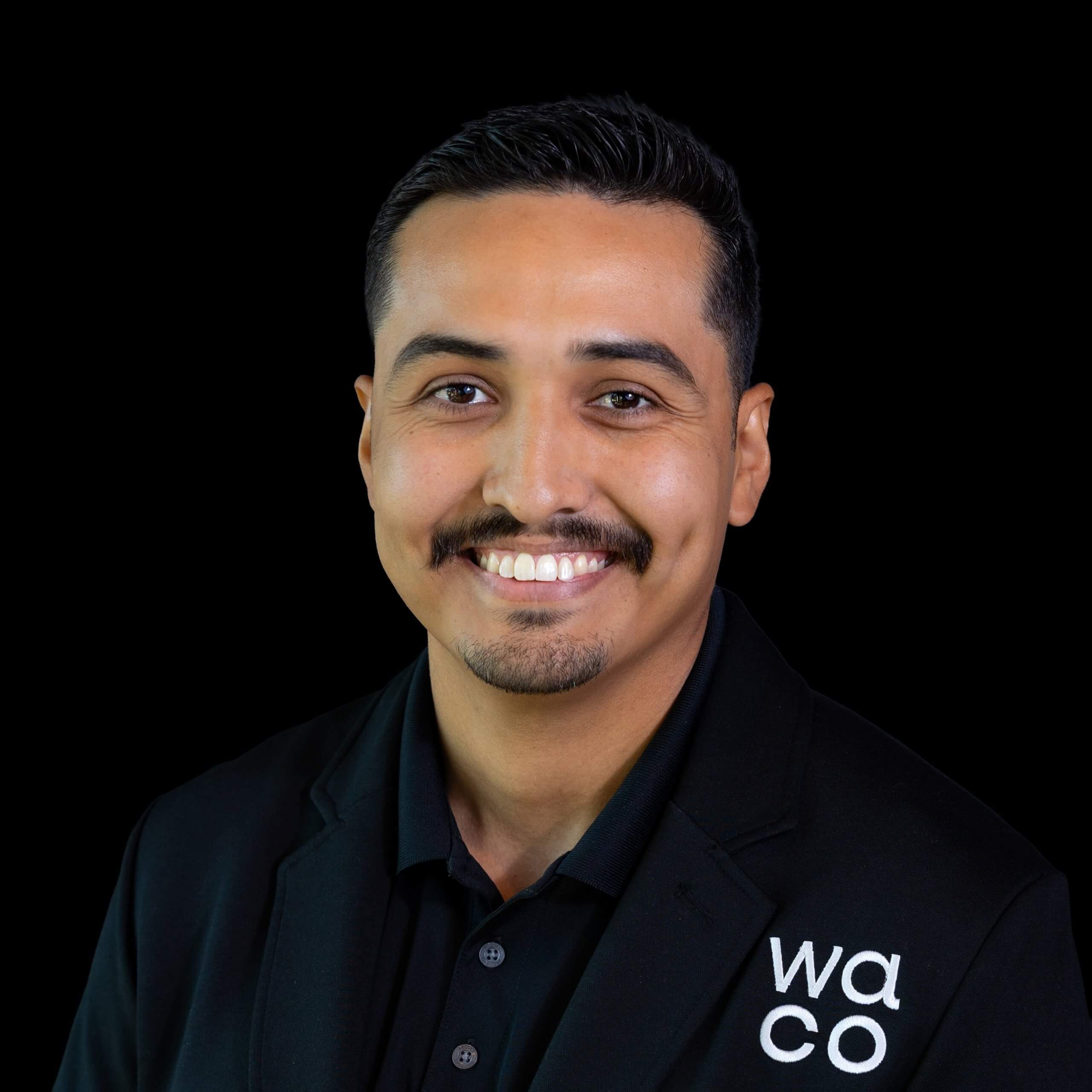 A smiling man with a mustache wearing a black blazer against a black background in McLennan County, Texas.