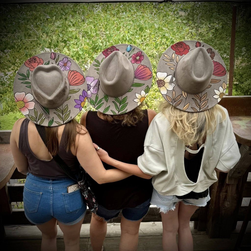 Three women from behind wearing matching floral-painted wide-brimmed hats while standing together in McLennan County, Texas.