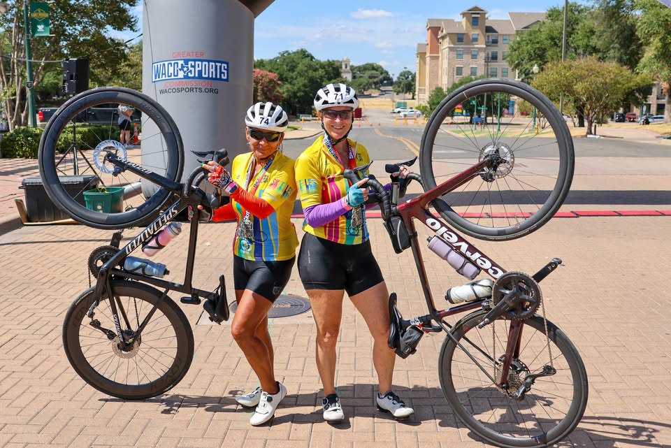 Two female cyclists in colorful jerseys pose holding their bicycles above their heads in McLennan County, Texas.