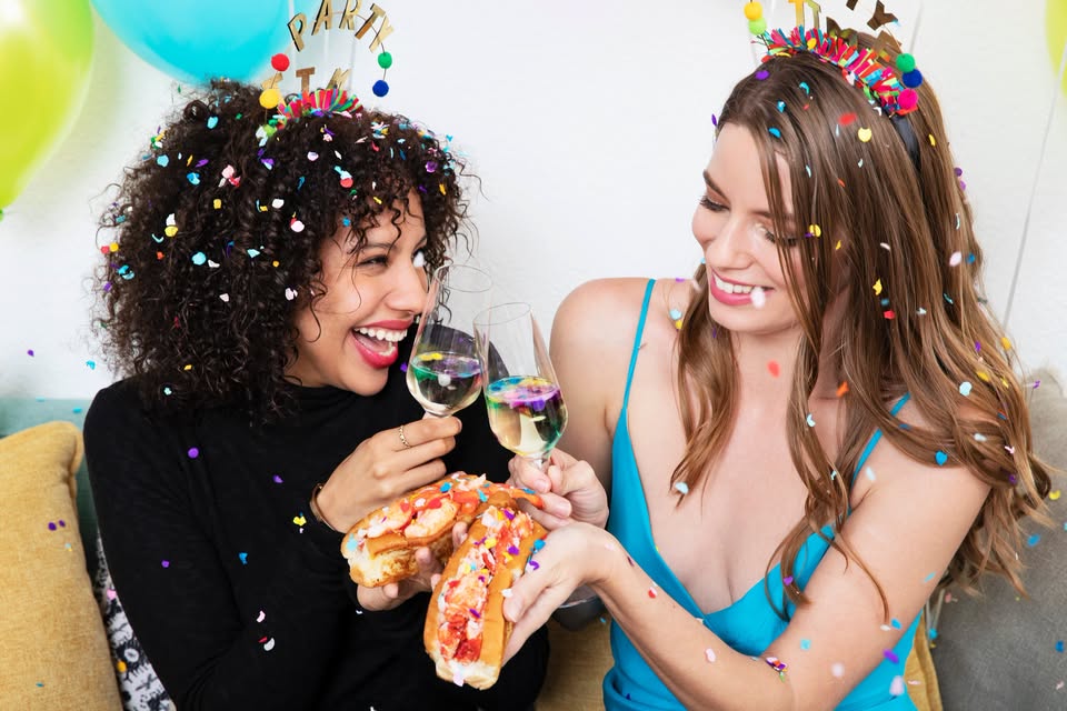 Two women wearing party headbands celebrate with champagne and lobster rolls amidst falling confetti in McLennan County, Texas.